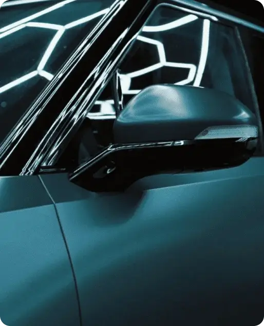 Tinted driver side window and door mirror on a dark teal car reflecting overhead shop lighting